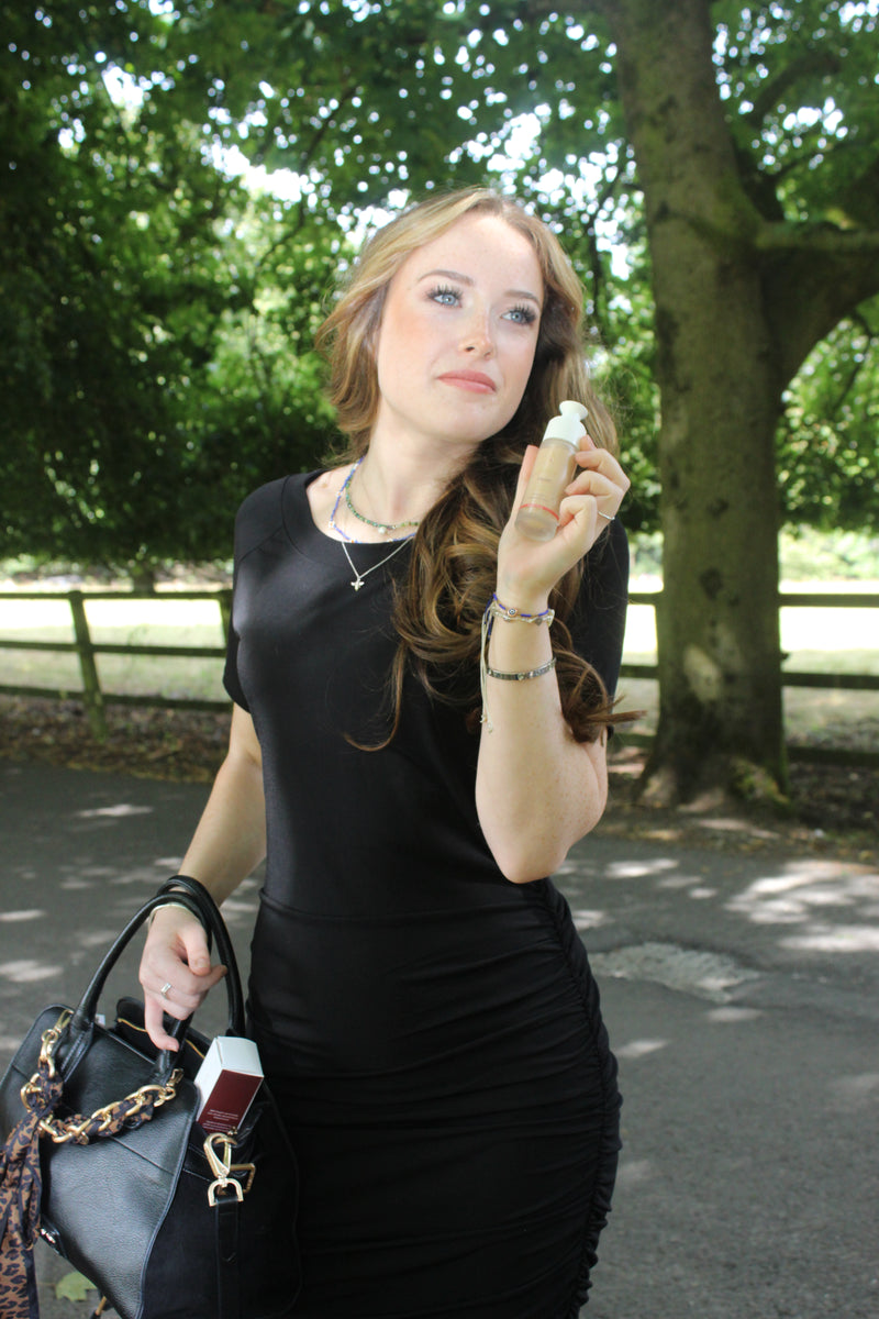 A woman in a black dress stands outdoors in a park, holding a small bottle to her nose as if smelling it. She is also holding a black handbag with a gold chain strap.
