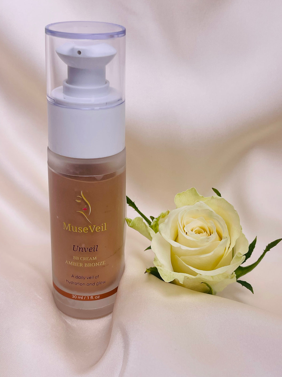 MuseVeil Unveil Airbrush Bronzer in a frosted pump bottle, styled next to a pale yellow rose on a draped cream satin background.