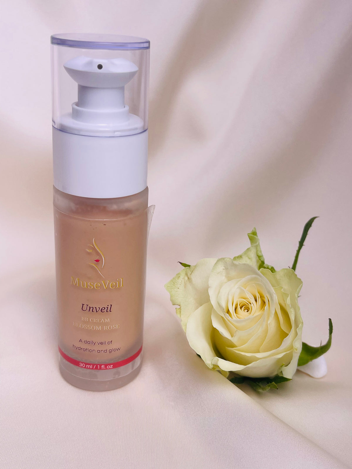 MuseVeil Unveil Hydrating Face Primer in a clear pump bottle next to a white rose on a draped cream fabric background.