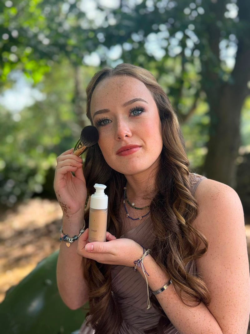 A woman outdoors holds a pump bottle of liquid foundation and a makeup brush, applying product to her face. The background shows blurred green foliage.