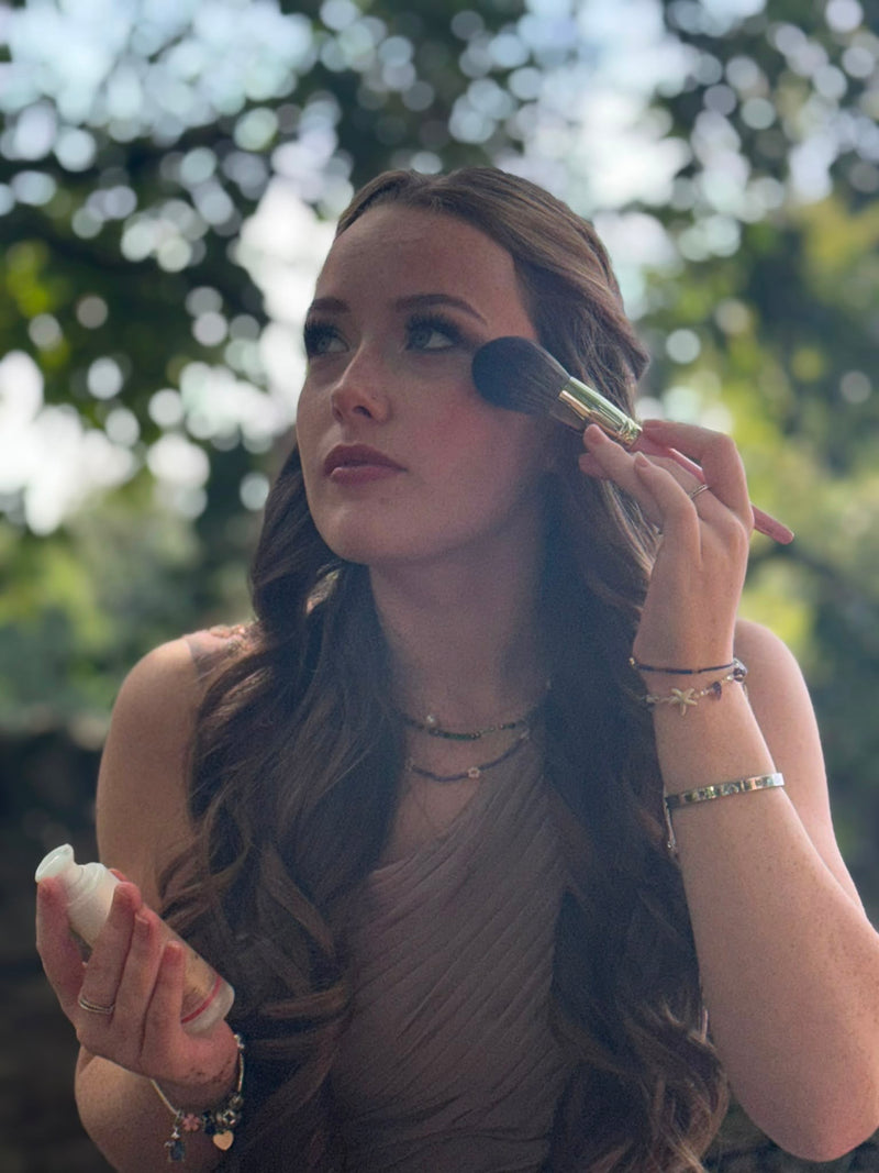 A woman with long wavy brown hair applies makeup with a brush outdoors, wearing a sleeveless taupe dress and layered necklaces, with a blurred green bokeh background.